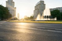 the sun is shining over a street and water feature fountain in the background of a big building