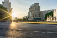 the sun is shining over a street and water feature fountain in the background of a big building