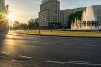 the sun is shining over a street and water feature fountain in the background of a big building