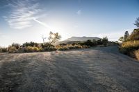 the sun is shining on a road with gravel and bushes in the foreground and mountains