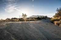 the sun is shining on a road with gravel and bushes in the foreground and mountains