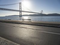 the sun shining over the water and a bridge across from it with benches on a curb beside it