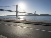 the sun shining over the water and a bridge across from it with benches on a curb beside it