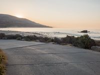 a person riding a surf board down the side of a road near the ocean at sunset