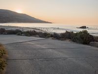a person riding a surf board down the side of a road near the ocean at sunset