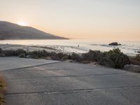 a person riding a surf board down the side of a road near the ocean at sunset