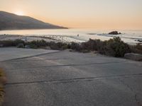 a person riding a surf board down the side of a road near the ocean at sunset