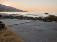a person riding a surf board down the side of a road near the ocean at sunset