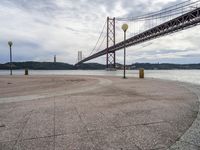 the suspension bridge is near the water in front of a building and mountain ranges and blue sky