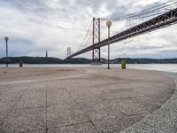 the suspension bridge is near the water in front of a building and mountain ranges and blue sky