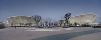 an empty circular plaza in front of tall buildings at dusk time, with people walking by, and two trees by the ground