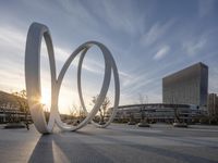 an outdoor sculpture outside in front of the buildings with sun setting behind it, in the city