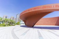 a building with multiple curved red concrete pillars in a garden area with several trees and skyscrapers around it