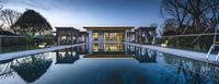 an elegant swimming pool lit up with lights at dusk in front of modern residence building