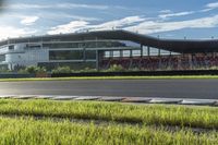 a car is driving down a paved road through the grass past a stadium building with the sun shining