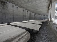 a very long sidewalk that is by some concrete buildings with a roof over it that has concrete walls