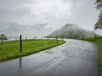 a road with a fence in a rainy day, with grass and mountains in the distance