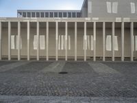 some columns outside a building with buildings on the other side of them, and sun shining over them