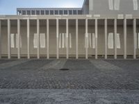 some columns outside a building with buildings on the other side of them, and sun shining over them