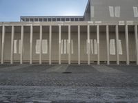 some columns outside a building with buildings on the other side of them, and sun shining over them