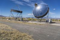 two large metal telescopes in a desert area near water and a building and some grass