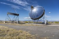 two large metal telescopes in a desert area near water and a building and some grass