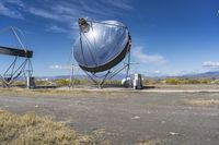 two large metal telescopes in a desert area near water and a building and some grass