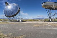 two large metal telescopes in a desert area near water and a building and some grass