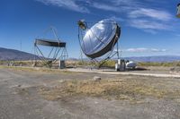 two large metal telescopes in a desert area near water and a building and some grass