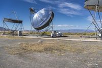 two large metal telescopes in a desert area near water and a building and some grass