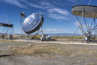 two large metal telescopes in a desert area near water and a building and some grass