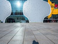 the shadow of someone on an empty tiled floor with a building in the background and large white clouds