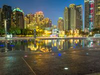 an empty sidewalk next to a large pond and tall buildings in the city skyline at night