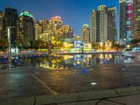 an empty sidewalk next to a large pond and tall buildings in the city skyline at night