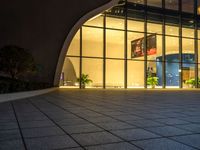 a large building is lit up at night with a glass facade and many potted plants in front