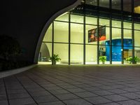 a large building is lit up at night with a glass facade and many potted plants in front