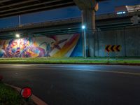 an underpass bridge next to a street at night with colorful murals on it, with a traffic sign next to the road