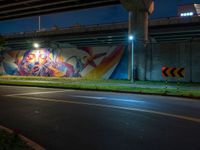 an underpass bridge next to a street at night with colorful murals on it, with a traffic sign next to the road