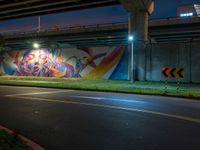 an underpass bridge next to a street at night with colorful murals on it, with a traffic sign next to the road