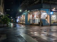 an outdoor cafe sits on a city street at night time with rain falling from the windows