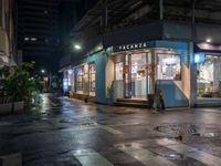 an outdoor cafe sits on a city street at night time with rain falling from the windows