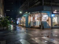 an outdoor cafe sits on a city street at night time with rain falling from the windows