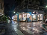 an outdoor cafe sits on a city street at night time with rain falling from the windows