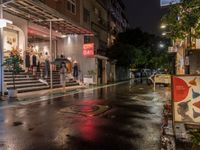 rainy, wet street lined with shops and people waiting at a curb in front of a building