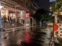 rainy, wet street lined with shops and people waiting at a curb in front of a building