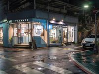 a store is on the corner of a wet street at night with a car passing by