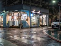 a store is on the corner of a wet street at night with a car passing by