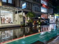 a wet sidewalk with a bunch of different storefronts on it at night,