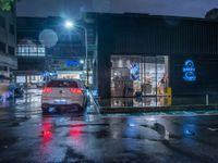 a small car parked at the street corner in front of a store front in rain at night