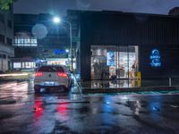 a small car parked at the street corner in front of a store front in rain at night